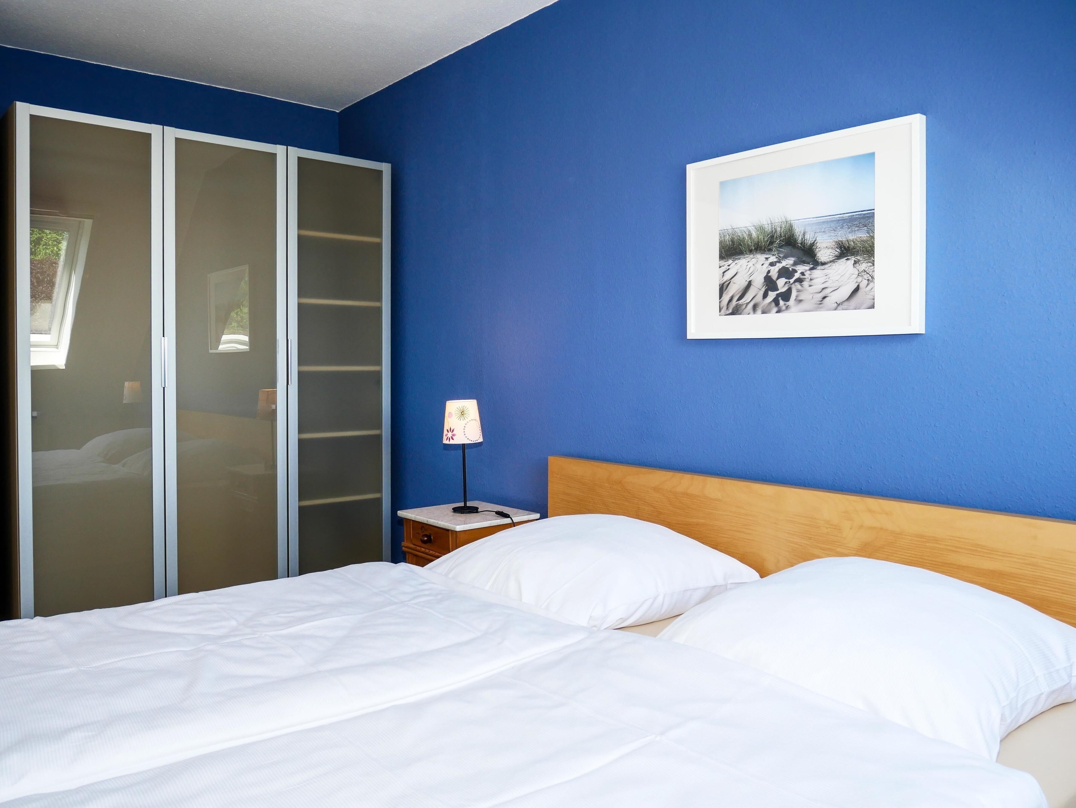 Ein Schlafzimmer mit Bett, weißer Bettwäsche, einem Nachttisch mit Lampe, einem gerahmten Bild an der Wand und einem Schiebetürenschrank.