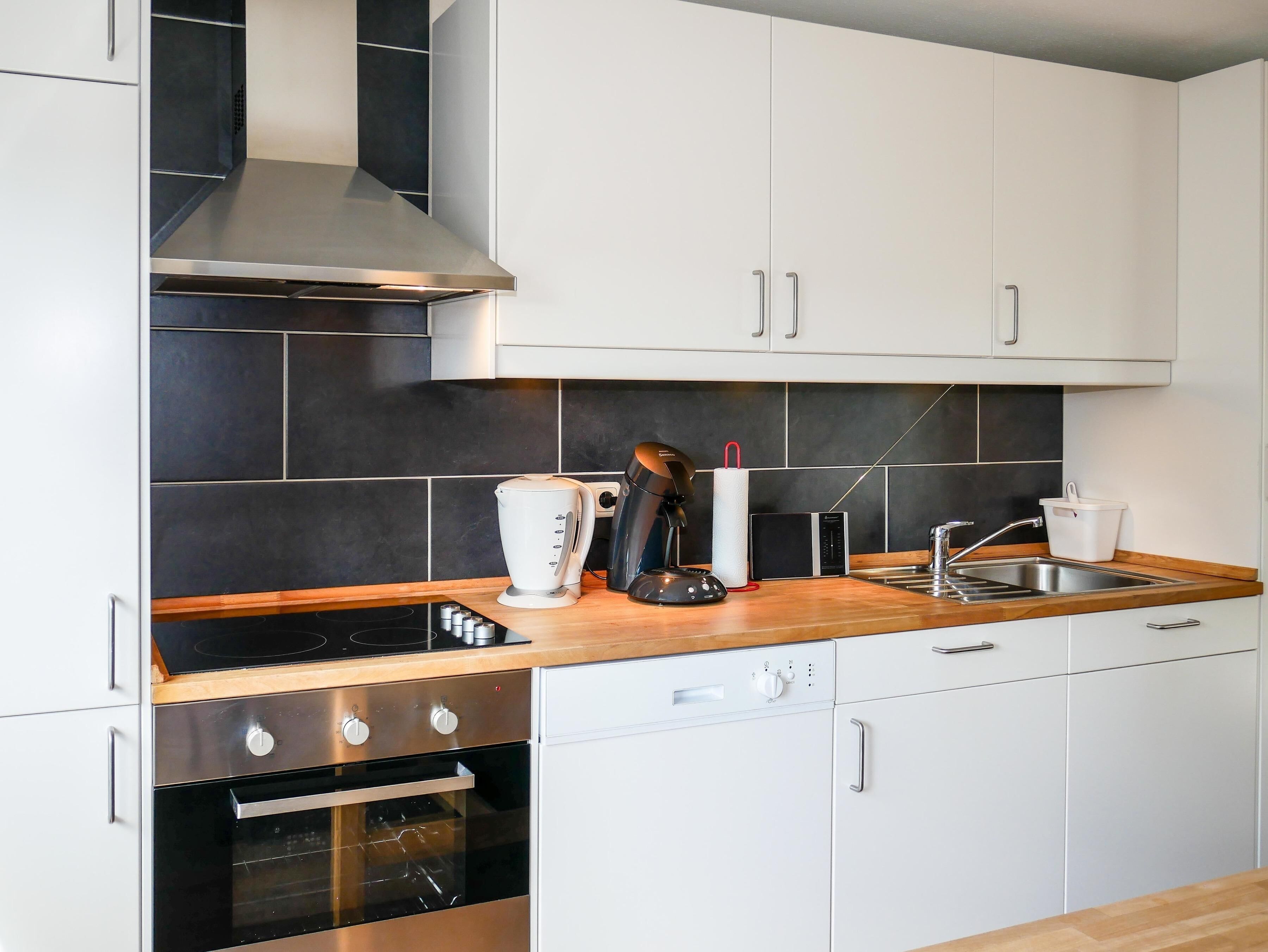 Eine moderne Küche mit weißen Schränken, einer schwarzen Küchenrückwand, einer Edelstahl-Dunstabzugshaube, einer Kaffeemaschine, einem Wasserkocher, einem Toaster und einem Geschirrspüler.
