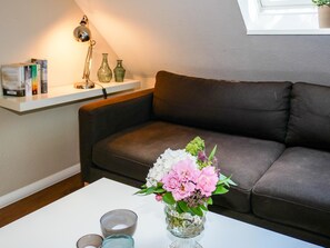 Ein Wohnzimmer mit einem braunen Sofa, einem weißen Couchtisch mit Blumen in einer Vase sowie einem Regal mit Büchern und Dekoration.