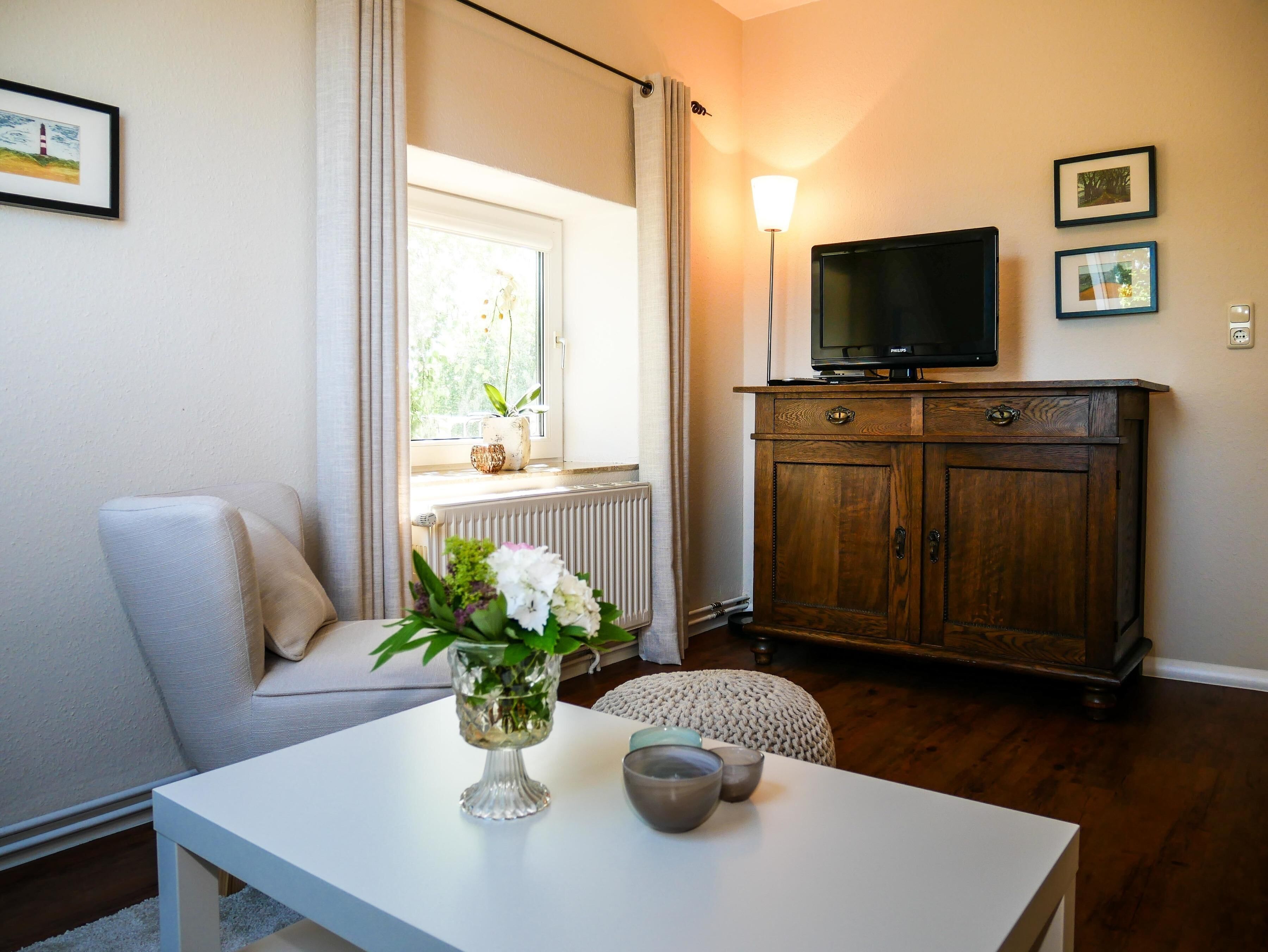 Ein Wohnzimmer mit einem weißen Sofa, einer Glasvase mit Blumen, einem Holzschrank und einem Fernseher auf einem Schrank.