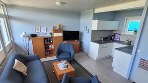 Un espacio habitable pequeño con sofá, dos sillones, una mesa de centro, televisor, estantería y kitchenette.
