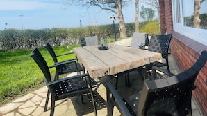 An outdoor patio with a wooden table and black metal chairs, surrounded by a brick wall and a view of a hedge and trees. - Vacation home Zur Alten Mühle - spacious living in Orth (Schwanitz) with garden & T (Fehmarn)