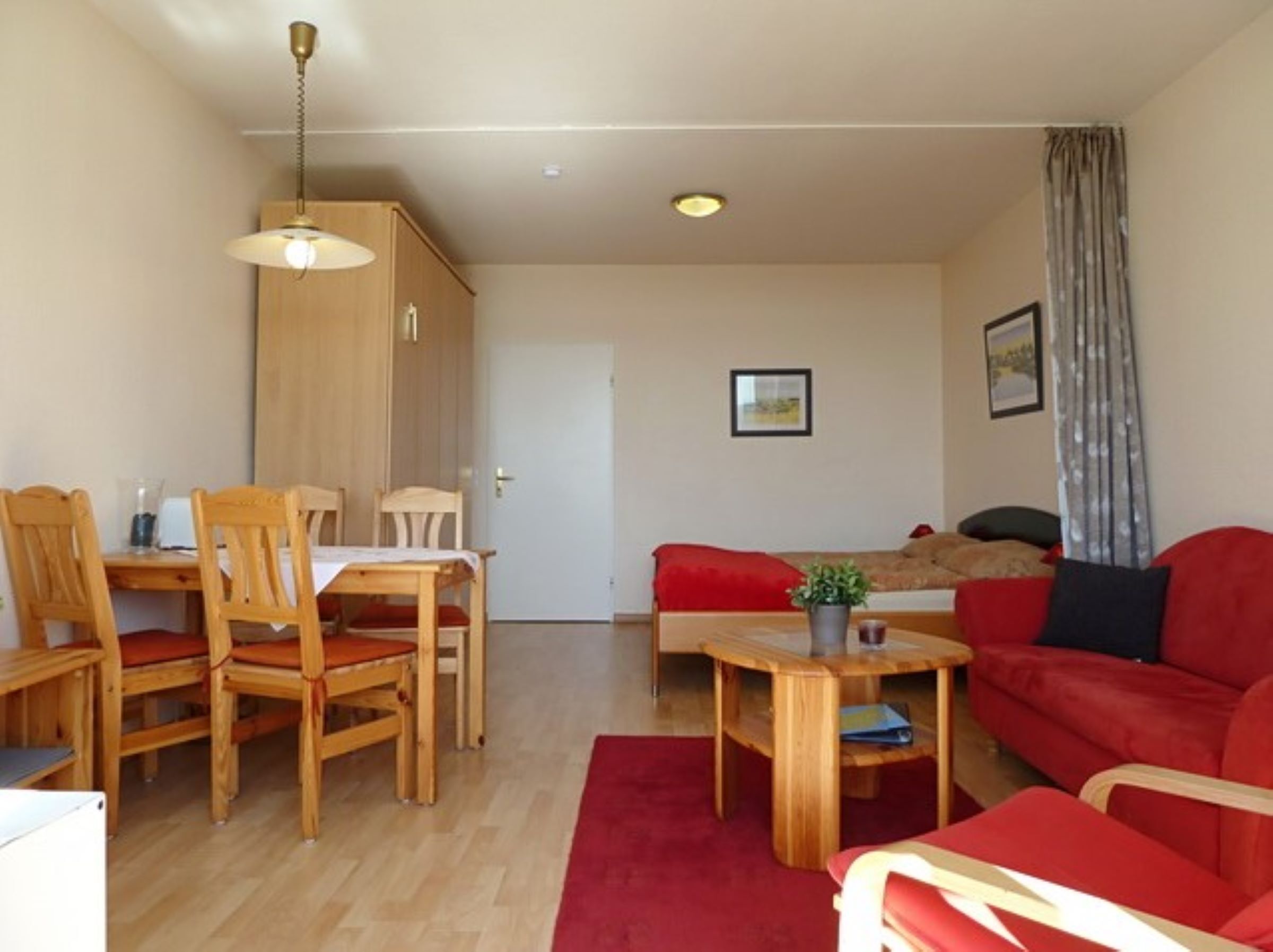Ein gemütliches Wohnzimmer mit einem roten Sofa, einem Holzespeisetisch und Stühlen. Ein kleiner Holztisch mit einer Pflanze und einem roten Teppich steht ebenfalls dort. Eine Tür führt in einen anderen Raum, und eine Deckenleuchte ist zu sehen.