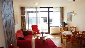 A hotel room with a red sofa, a dining table with chairs, a TV, and a balcony with a view. - Relax in the cozy vacation apartment on the Baltic Sea in Fehmarn! (Fehmarn)