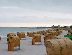 Fileiras de cadeiras de vime com números, dispostas numa praia de areia, com um corpo de água e um cais ao fundo.