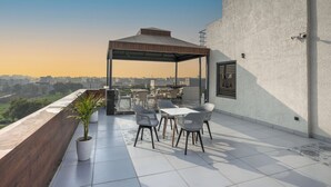 A rooftop terrace with a covered seating area, a potted plant, and a view of the cityscape. - Madhuram Inn (Gautam Buddh Nagar)