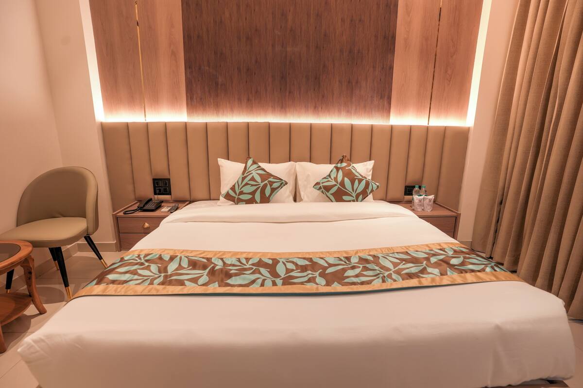 Habitación de hotel con una cama grande, una mesita de noche con un teléfono y una botella, una silla y un ventanal con cortinas.