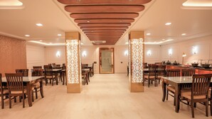 A spacious dining area with multiple tables and chairs, wooden ceiling panels, and decorative light fixtures. - Madhuram Inn (Gautam Buddh Nagar)