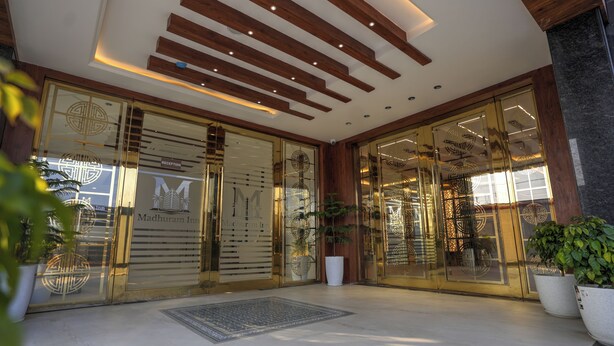 A hotel entrance with a reception desk, glass doors, and potted plants. - Madhuram Inn (Gautam Buddh Nagar)