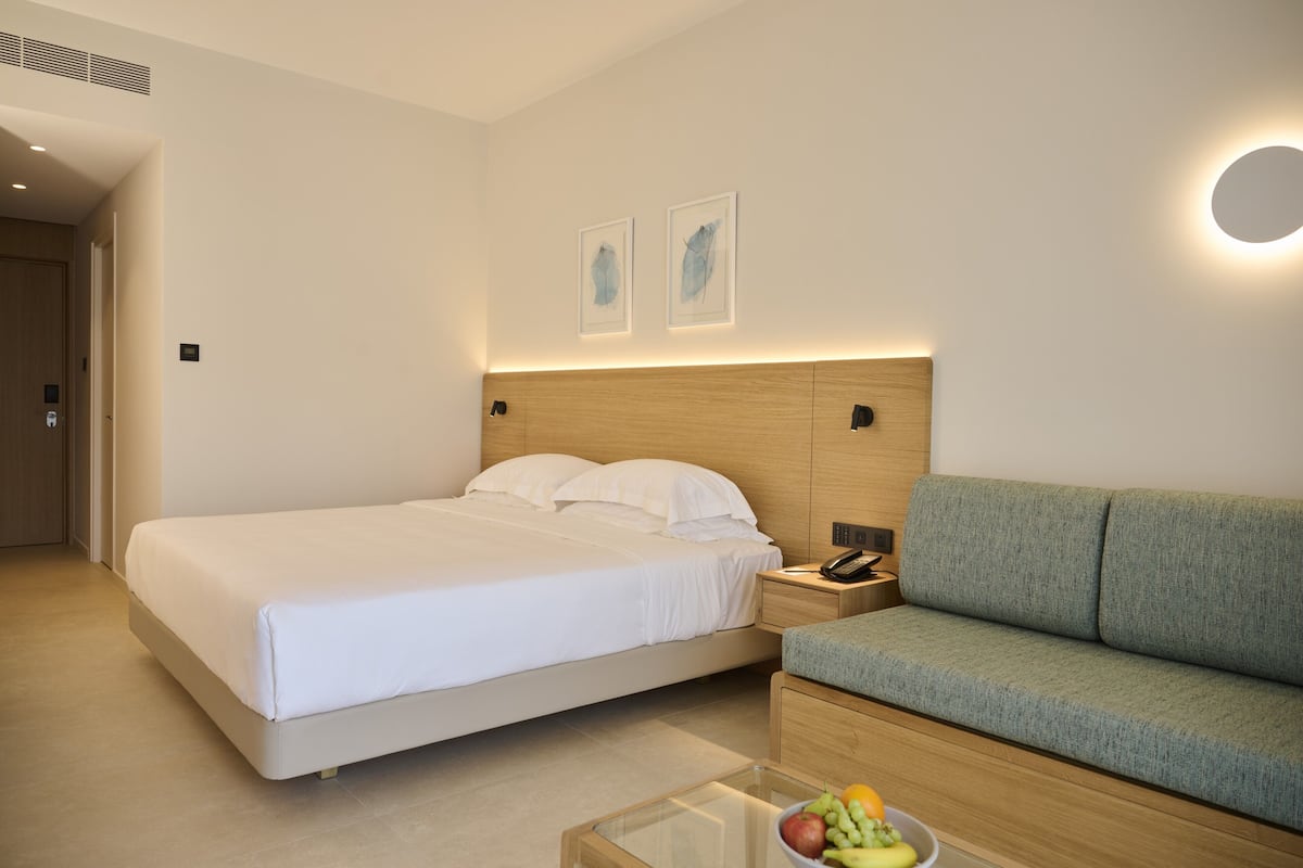 Una habitación de hotel con una cama, un sofá verde, una mesita de noche con un teléfono y una mesa de centro de cristal con fruta.