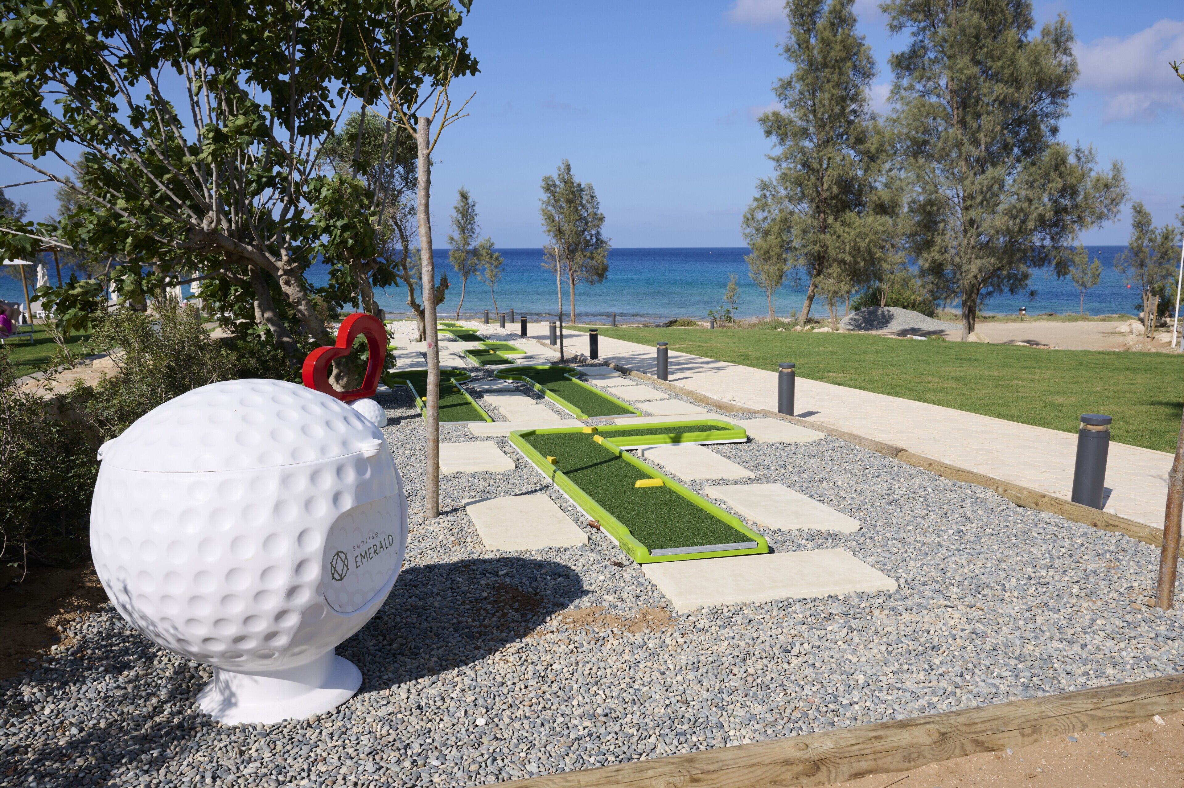 Un campo de minigolf con una pelota de golf blanca grande, césped artificial verde y una vista despejada del mar.