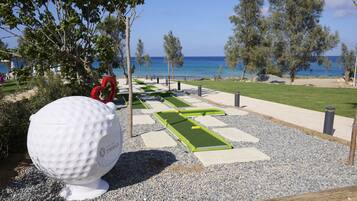 Un campo de minigolf con una pelota de golf blanca grande, césped artificial verde y una vista despejada del mar.