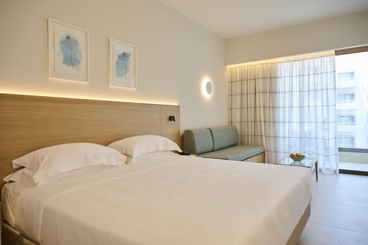 Habitación de hotel con una cama grande, un sofá, una mesita con un frutero y dos cuadros en la pared.