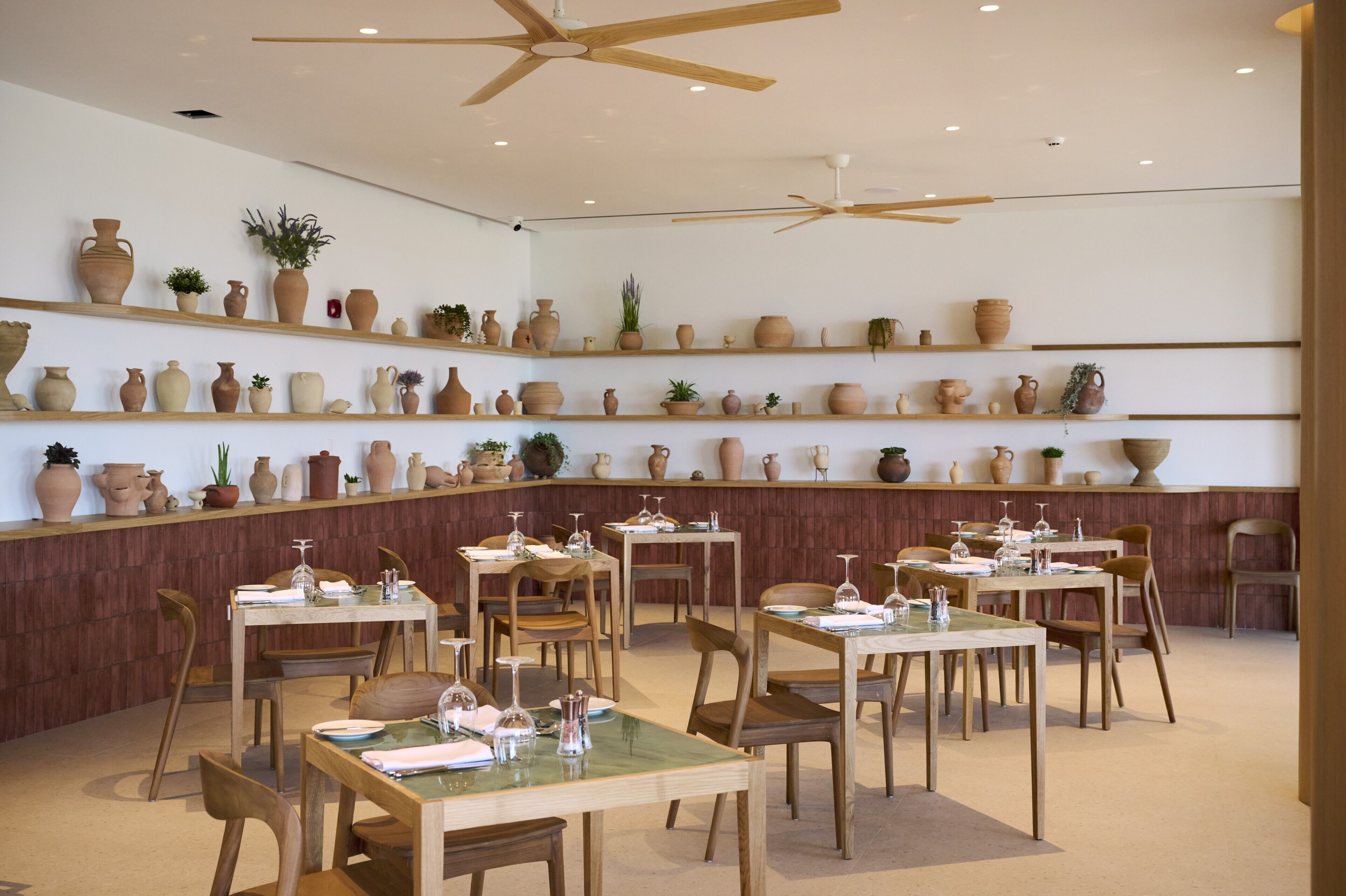 A restaurant with multiple tables and chairs, wooden ceiling fans, and shelves with various pottery and plants.