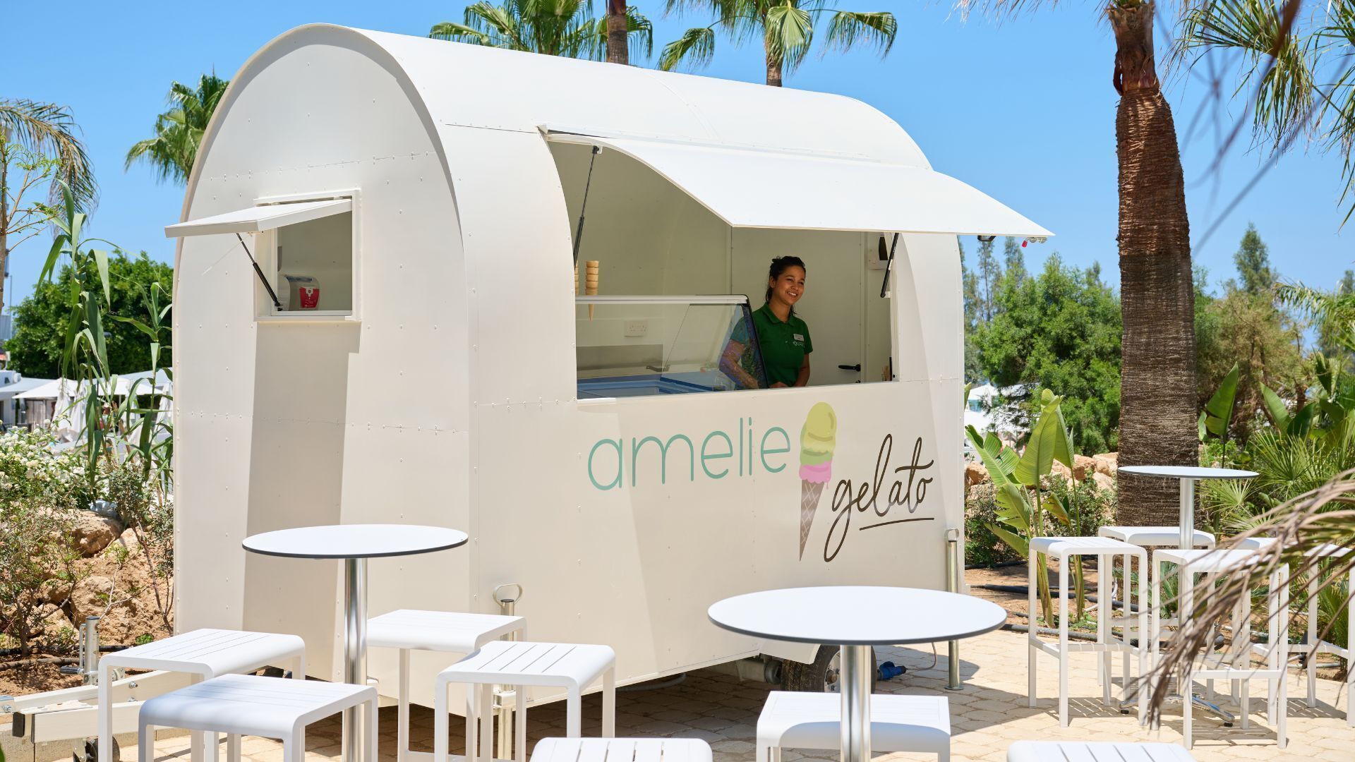 A small, white, curved structure with the name 'amelie gelato' on its side, a person inside, and outdoor seating with tables and chairs.