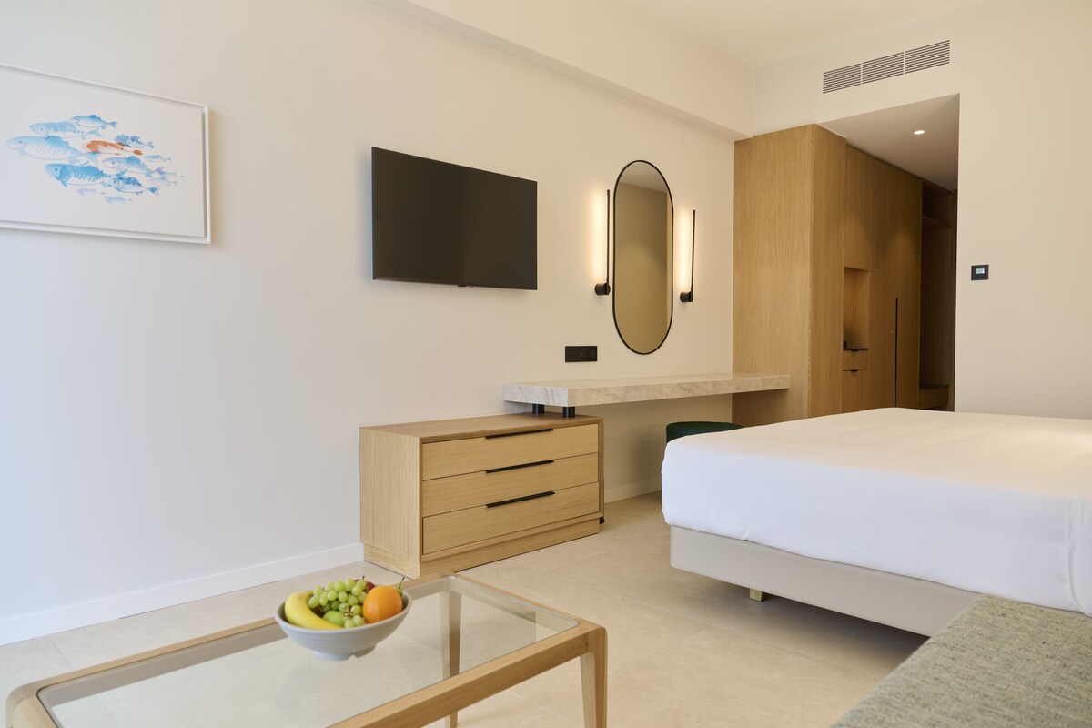 Una habitación de hotel moderna con una cama, un escritorio con cajones, un televisor de pantalla plana, un espejo y un frutero sobre una mesa de vidrio.