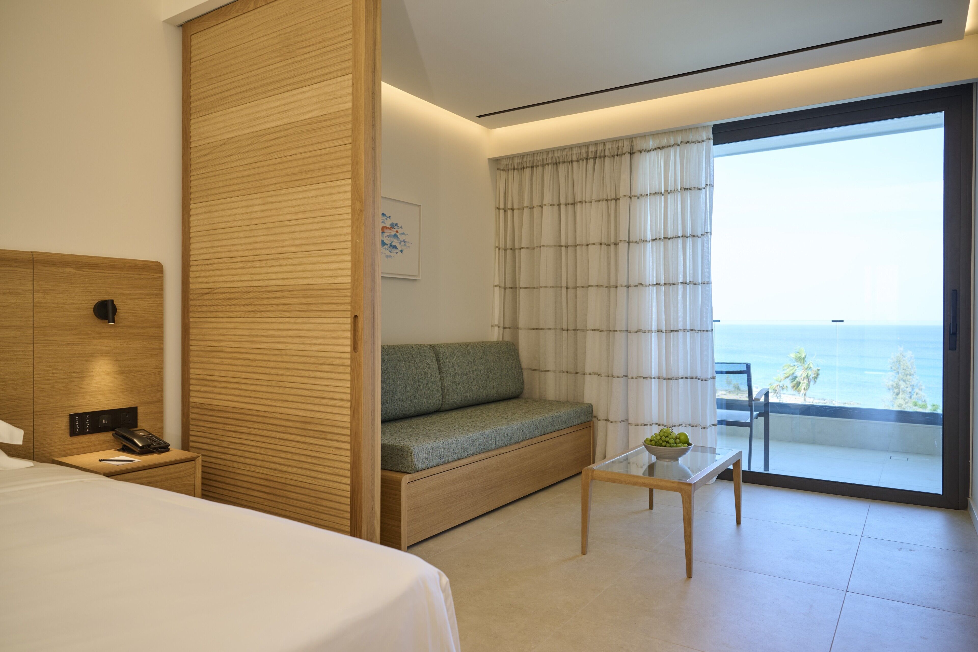 Una habitación de hotel moderna con una cama, un sofá, una mesita con una planta, un teléfono en una mesita de noche y vistas al océano a través de un ventanal grande.