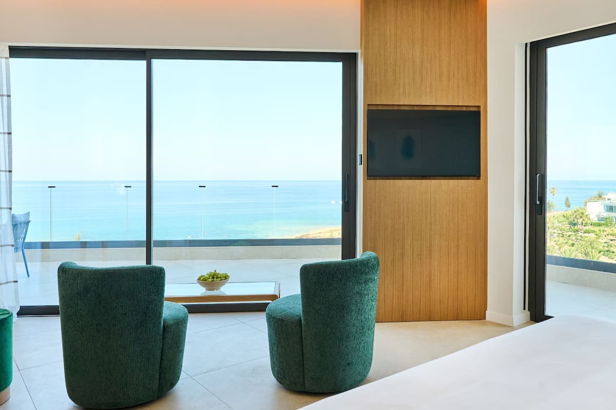 Una habitación moderna con un gran ventanal corredera que da a un balcón con vistas al mar, dos sillones tapizados en verde, una mesita con un cuenco de fruta y un televisor de pantalla plana montado en un panel de madera.