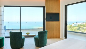 Ein modernes Zimmer mit einer großen SchiebeglasTür, die auf einen Balkon mit Meerblick führt, zwei grüne Polsterstühle, einem kleinen Tisch mit Obstschale und einem flachbildfernseher, der an einem Holzpaneel angebracht ist.