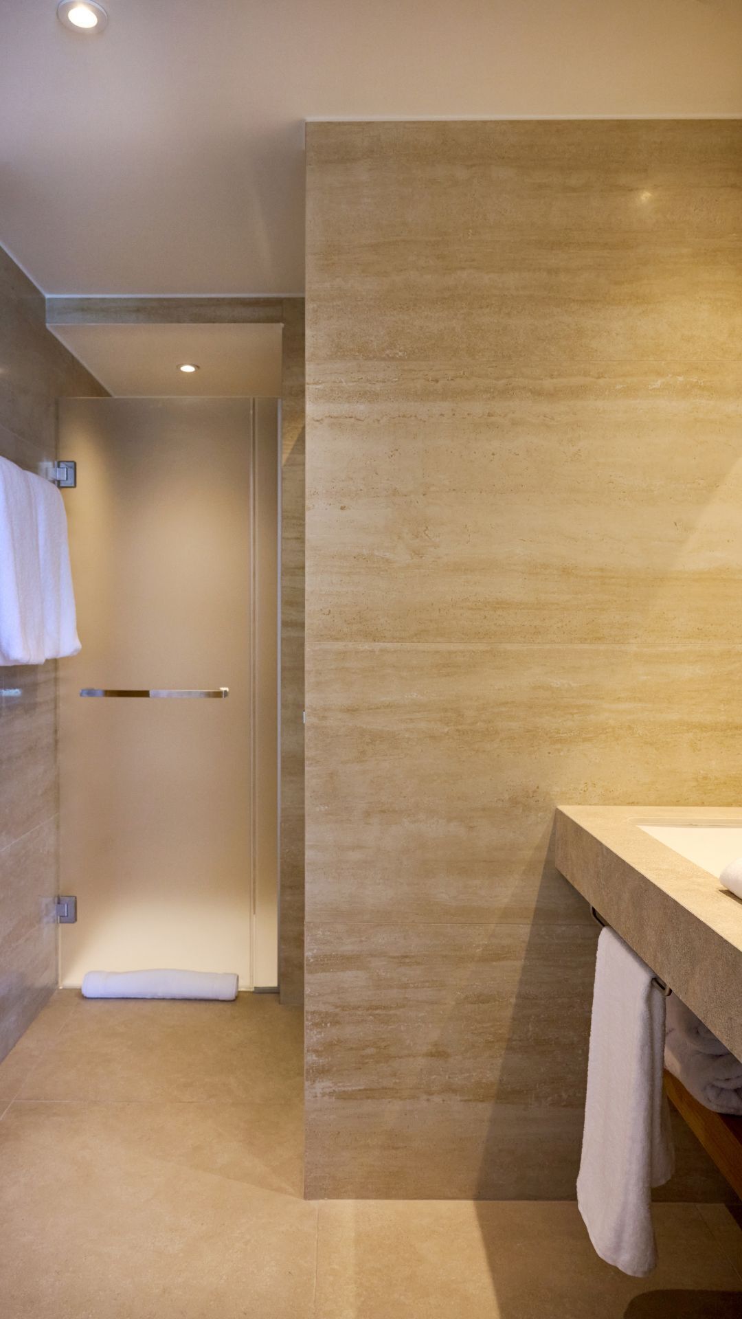 A modern bathroom with a beige tiled wall, a glass shower door, and a wooden vanity with a sink.