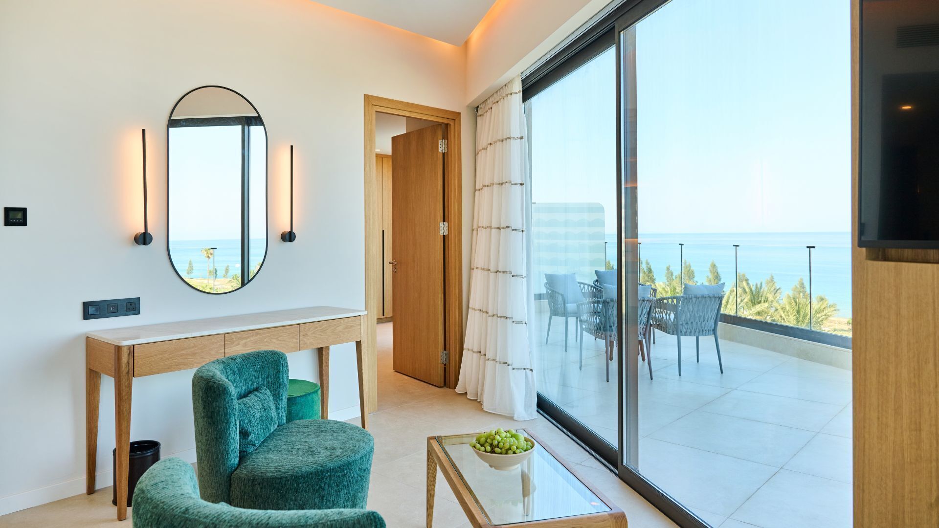 Una sala de estar moderna con un ventanal grande, un espejo redondo, una consola de madera, un sillón verde y una mesa de centro de vidrio.