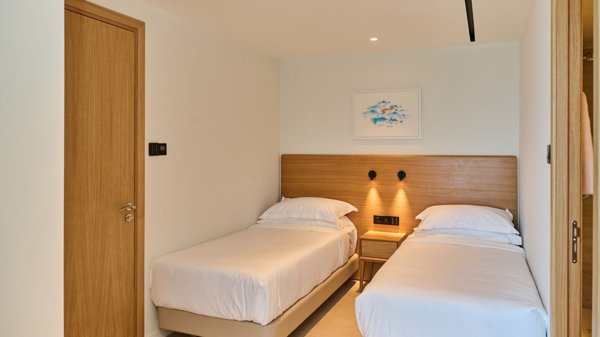 A small, well-lit bedroom with two single beds, a wooden headboard, a bedside table with a lamp, and a wall-mounted artwork.