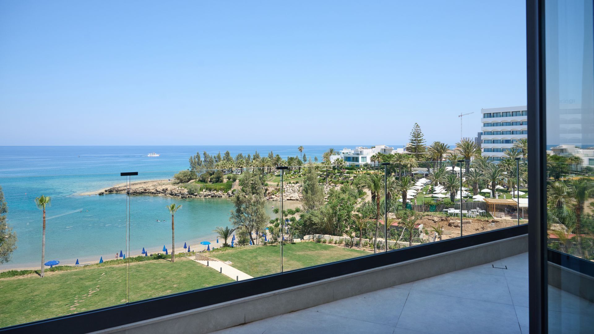 A view from a balcony overlooking a beach with a clear sky, a distant boat, and a resort area with palm trees and buildings.