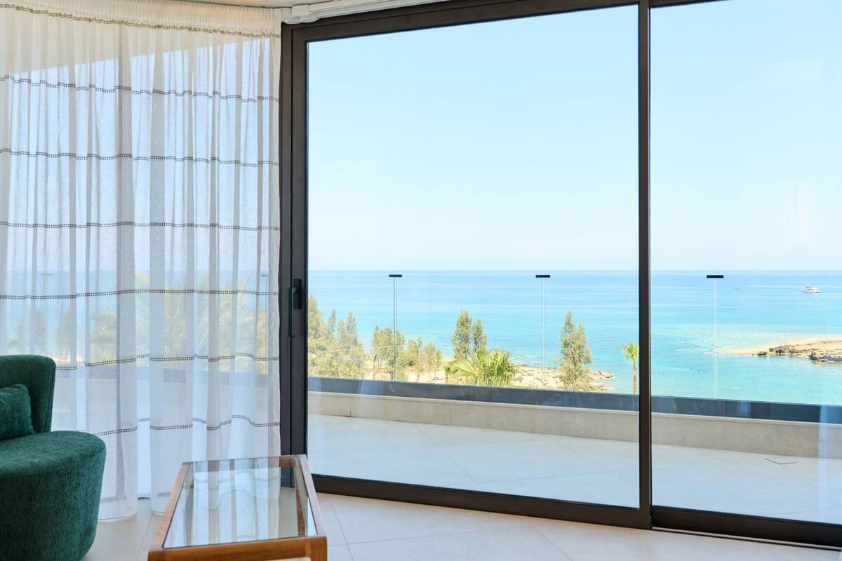 Una habitación con un gran ventanal corredera que da a un balcón con vistas al mar y un barco a lo lejos.