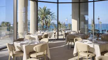 Un restaurant avec des tables dressées pour le service, de grandes fenêtres et une vue sur l’océan.