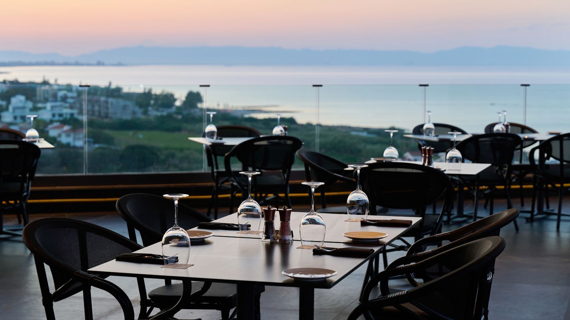 A restaurant with tables set for dining, featuring a view of a body of water and distant buildings.