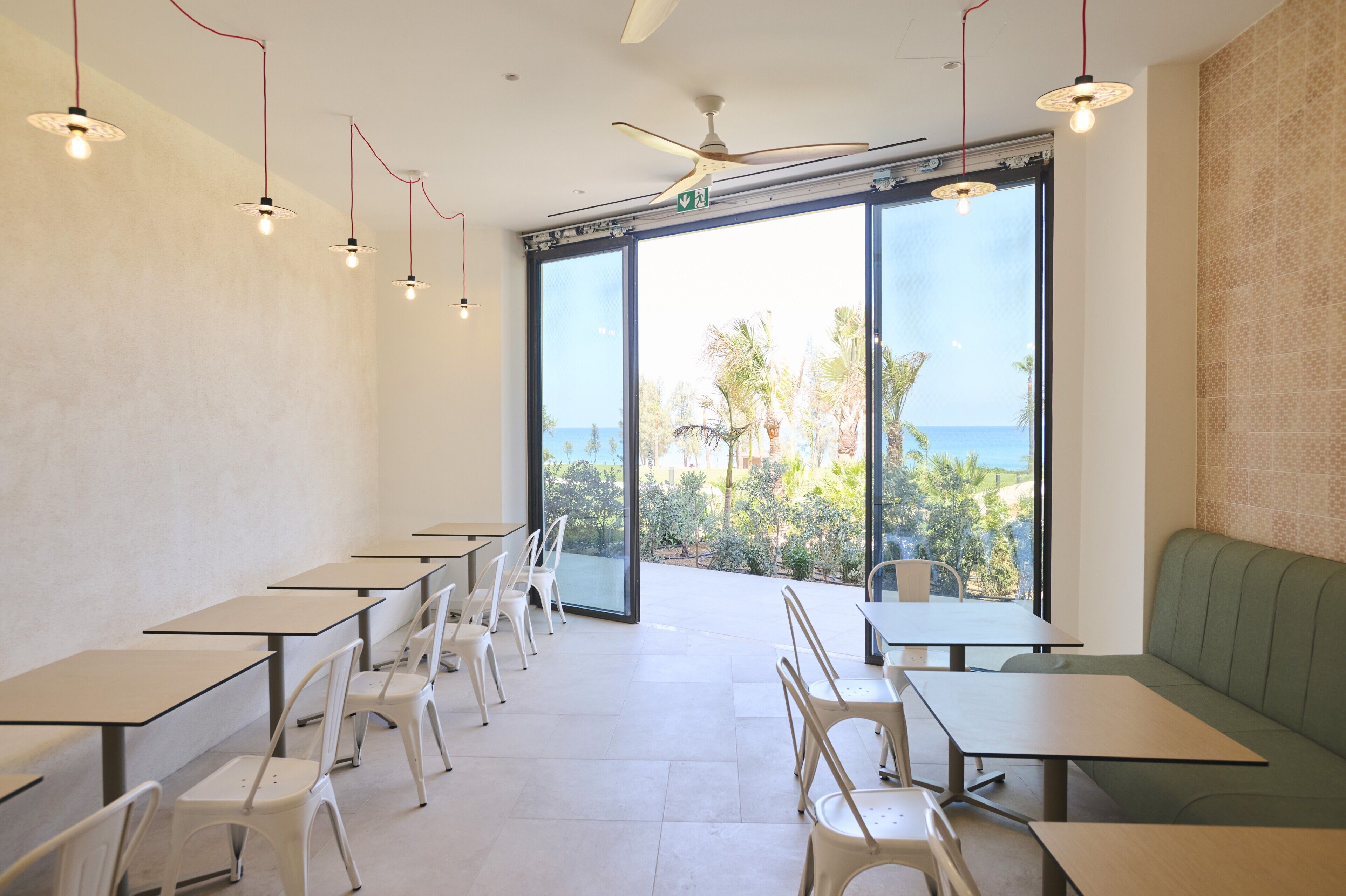 Un restaurant avec des tables et des chaises, de grandes fenêtres donnant sur des palmiers et l’océan, et un ventilateur de plafond.