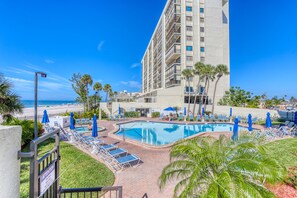 A hotel pool area with lounge chairs and umbrellas, a beach view, and a tall building in the background. - ☀Beachfront Condo🐬End Unit🍹Ocean Sands I-501🌴Heated Pool/Hot Tub🏖️Beach Views (Madeira Beach)