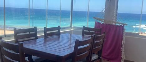 A wooden dining table with six chairs, large windows with a view of the ocean, and a beach visible through the windows.