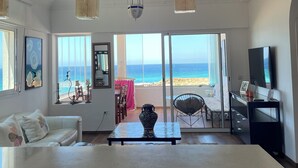 A spacious living room with a large window offering a view of the beach. There is a white sofa, a dining table, and a TV stand. - atico azul (Tangier)