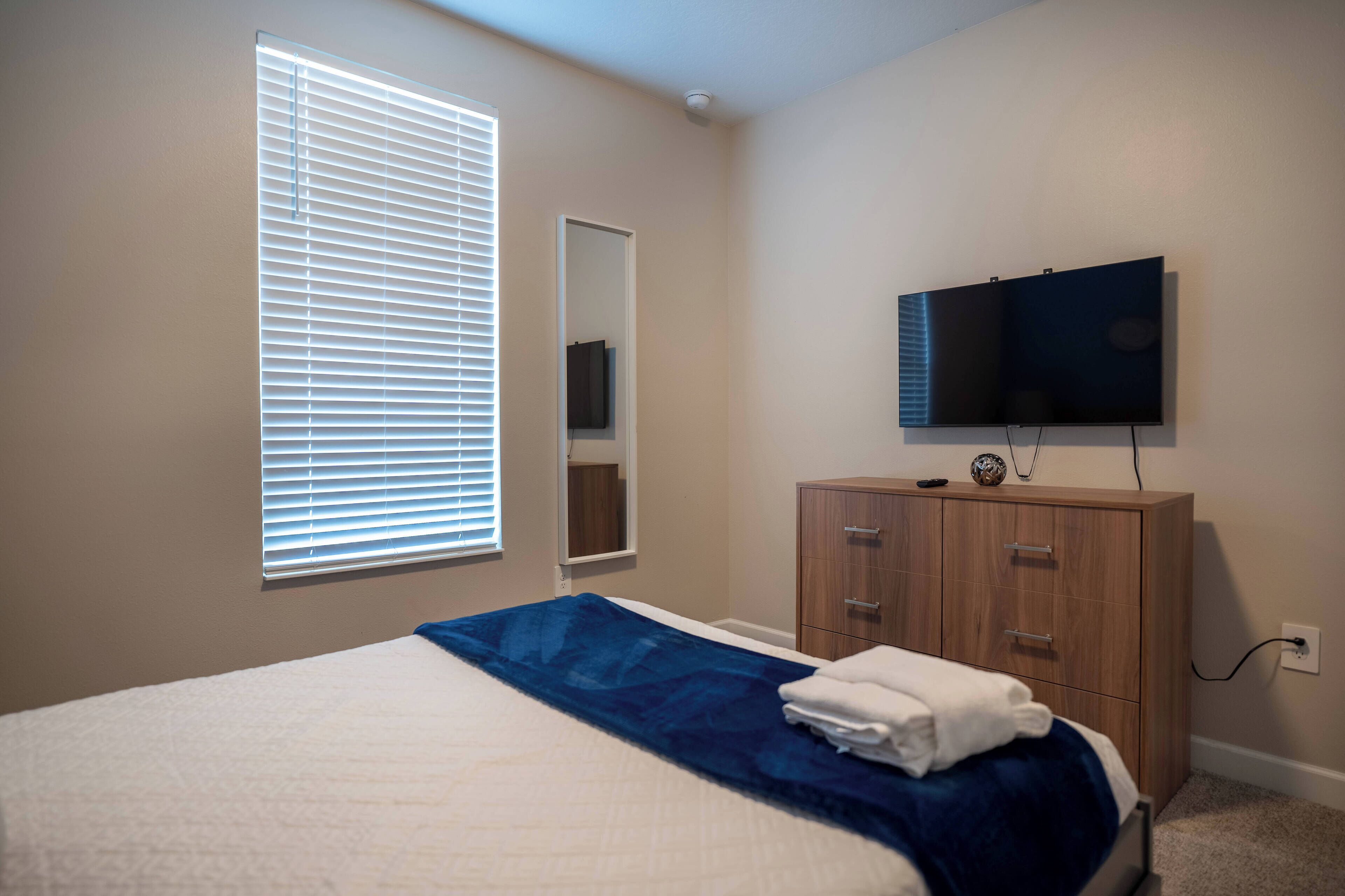 A bedroom with a bed, a television mounted on the wall, a wooden dresser, and a window with blinds.