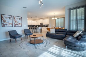 A modern living room with a sofa, chairs, and a coffee table. There is a kitchen in the background with a dining area. - 6BR Luxury Pool Home Near Disney Champions (Davenport)