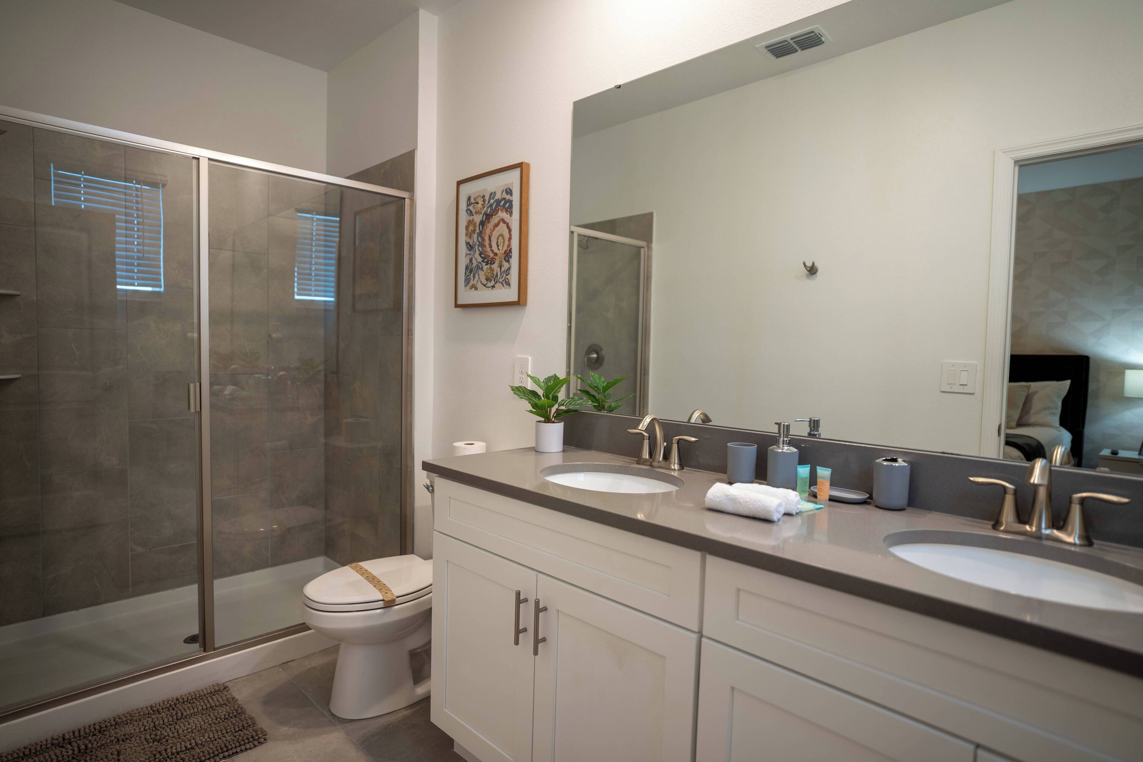 A modern bathroom with a glass-enclosed shower, a white toilet, and a vanity with two sinks and a large mirror.