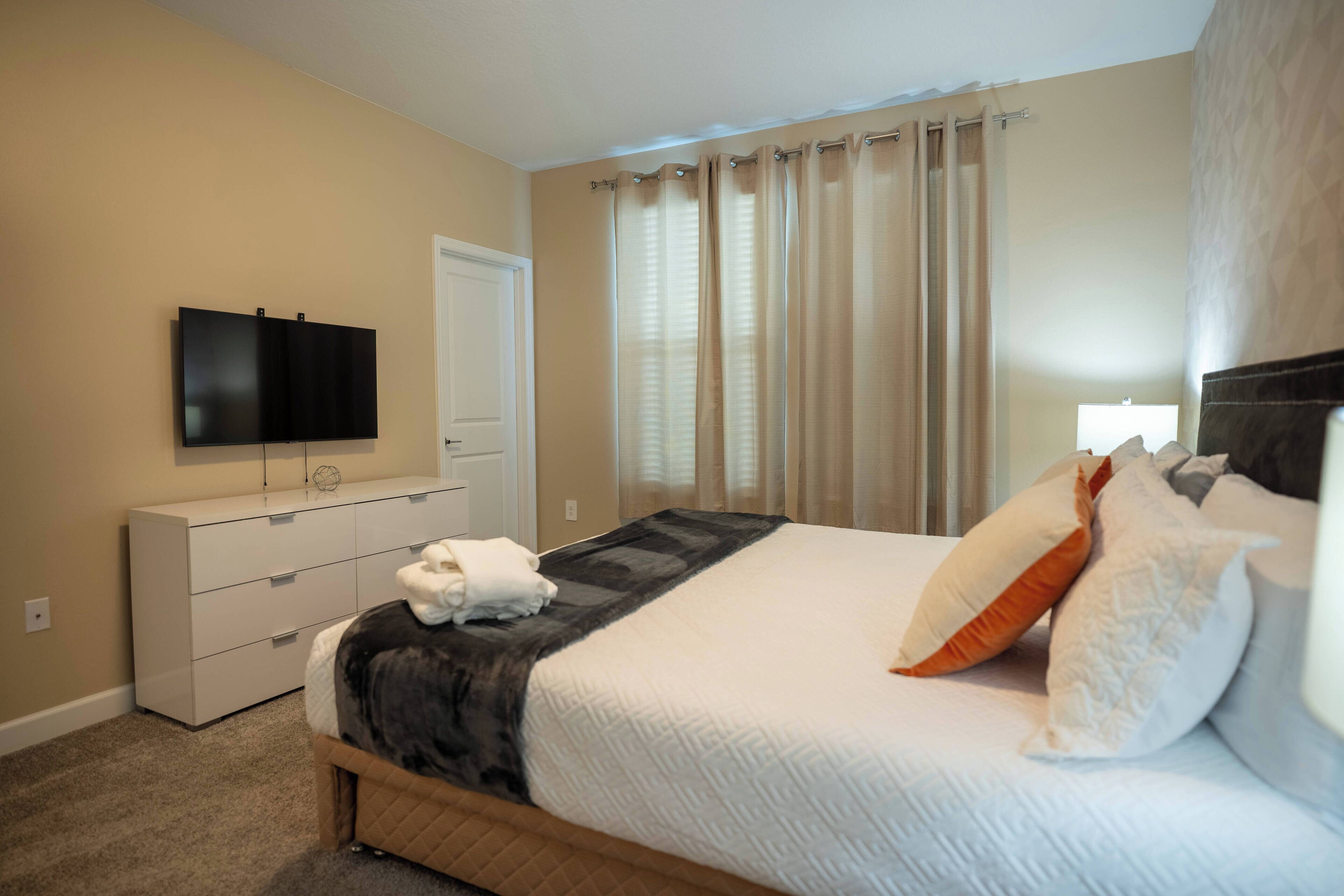A bedroom with a bed, a flat-screen TV, a dresser, and a window with curtains.