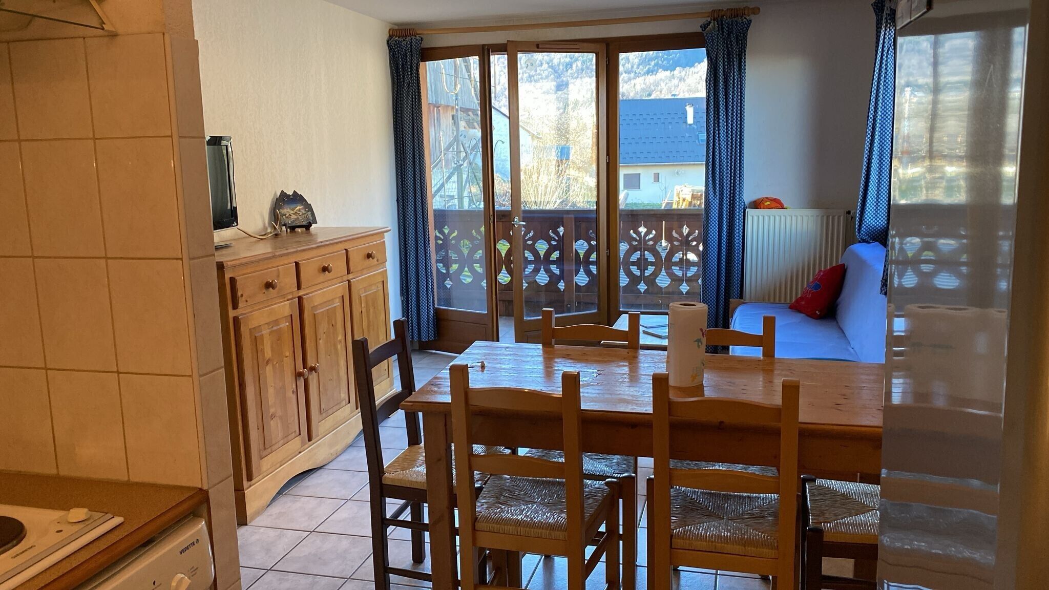 Ein Essbereich mit Tisch und Stühlen, eine Küche mit Schränken und Blick auf einen Balkon mit Geländer und ein Gebäude im Hintergrund.