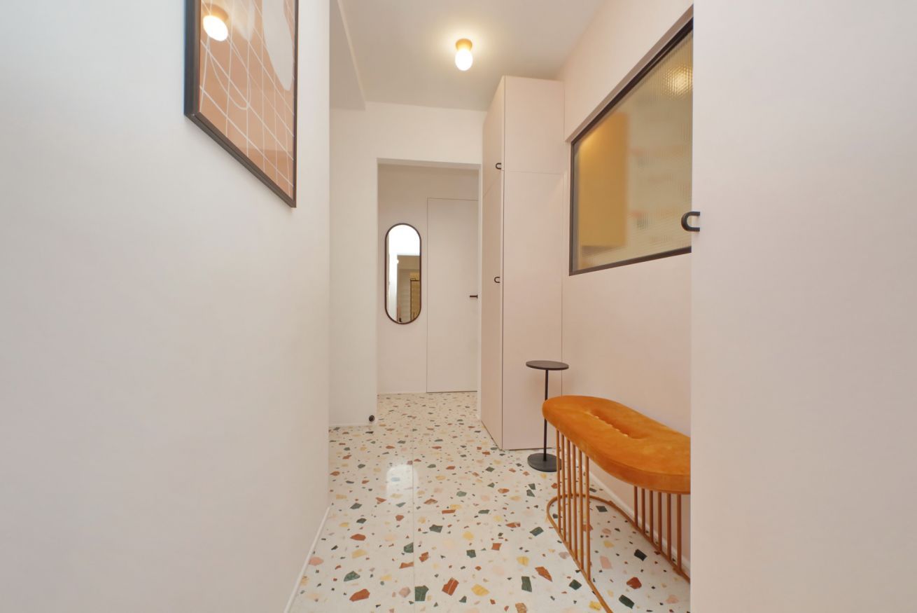 Un passadís estret amb sòl de terrazo, un mirall rodó a la paret i un petit tamboret.
