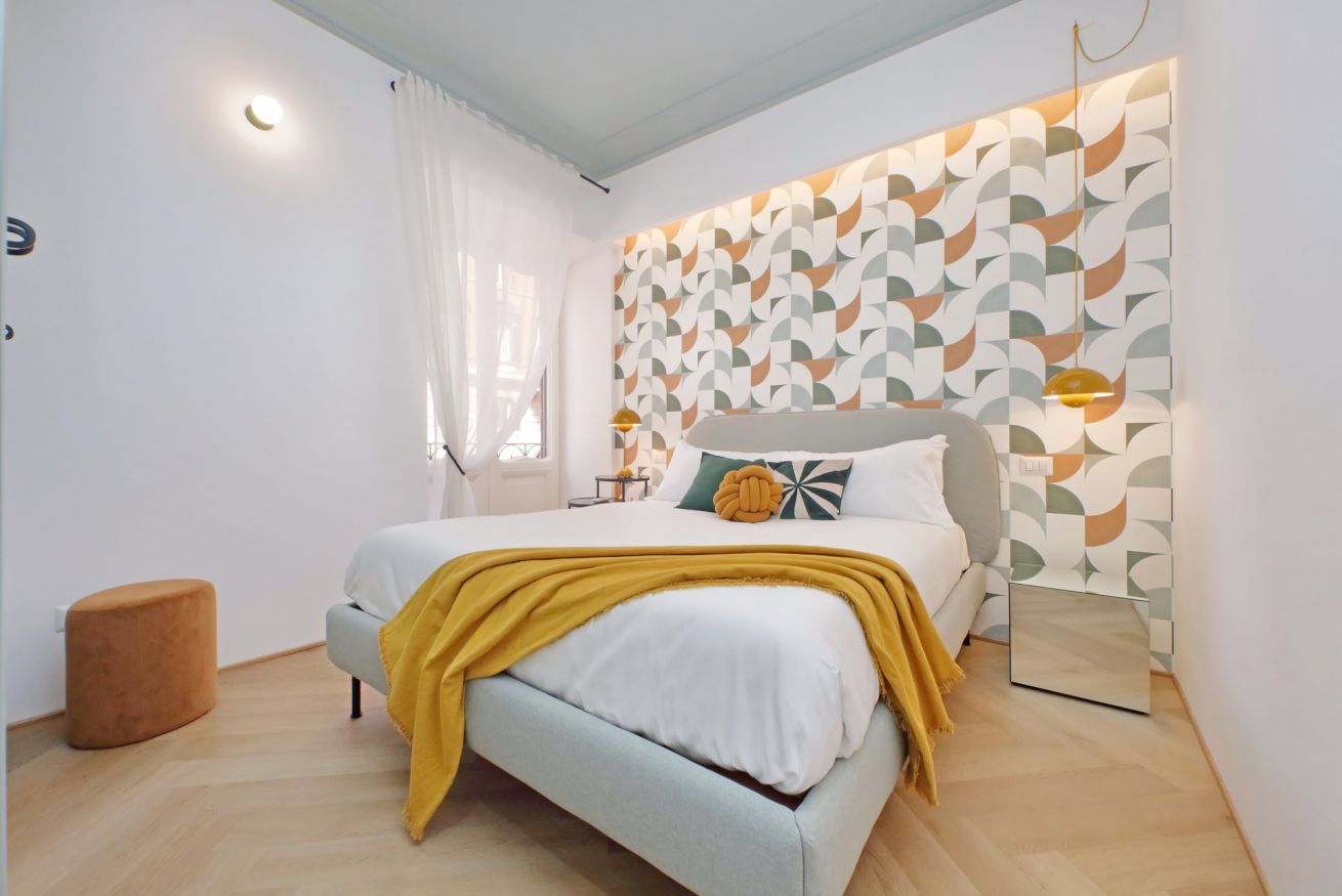 A modern bedroom with a bed, a bedside table, a mirror, and a patterned wall.