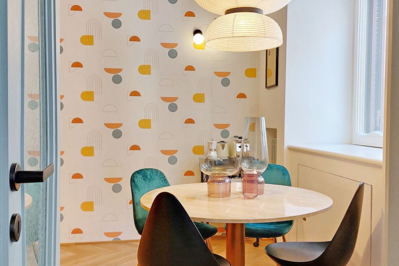 Un comedor con una mesa redonda, sillas color turquesa y un papel tapiz con motivos geométricos.