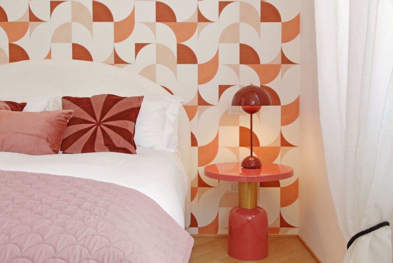 Une chambre à coucher avec un lit, des oreillers, une table ronde avec une lampe et un mur décoré de motifs.