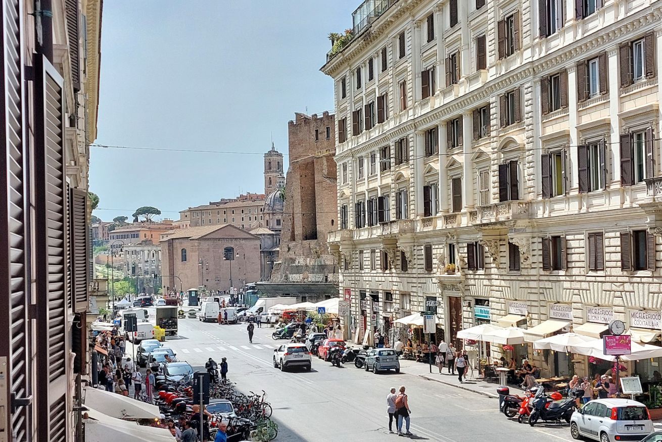 A bustling street with historic buildings, outdoor cafes, and a prominent tower in the background.