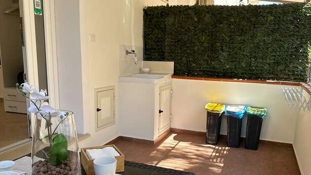 A balcony with a table, chairs, a glass container with pebbles, a white sink, and a green leafy wall.
