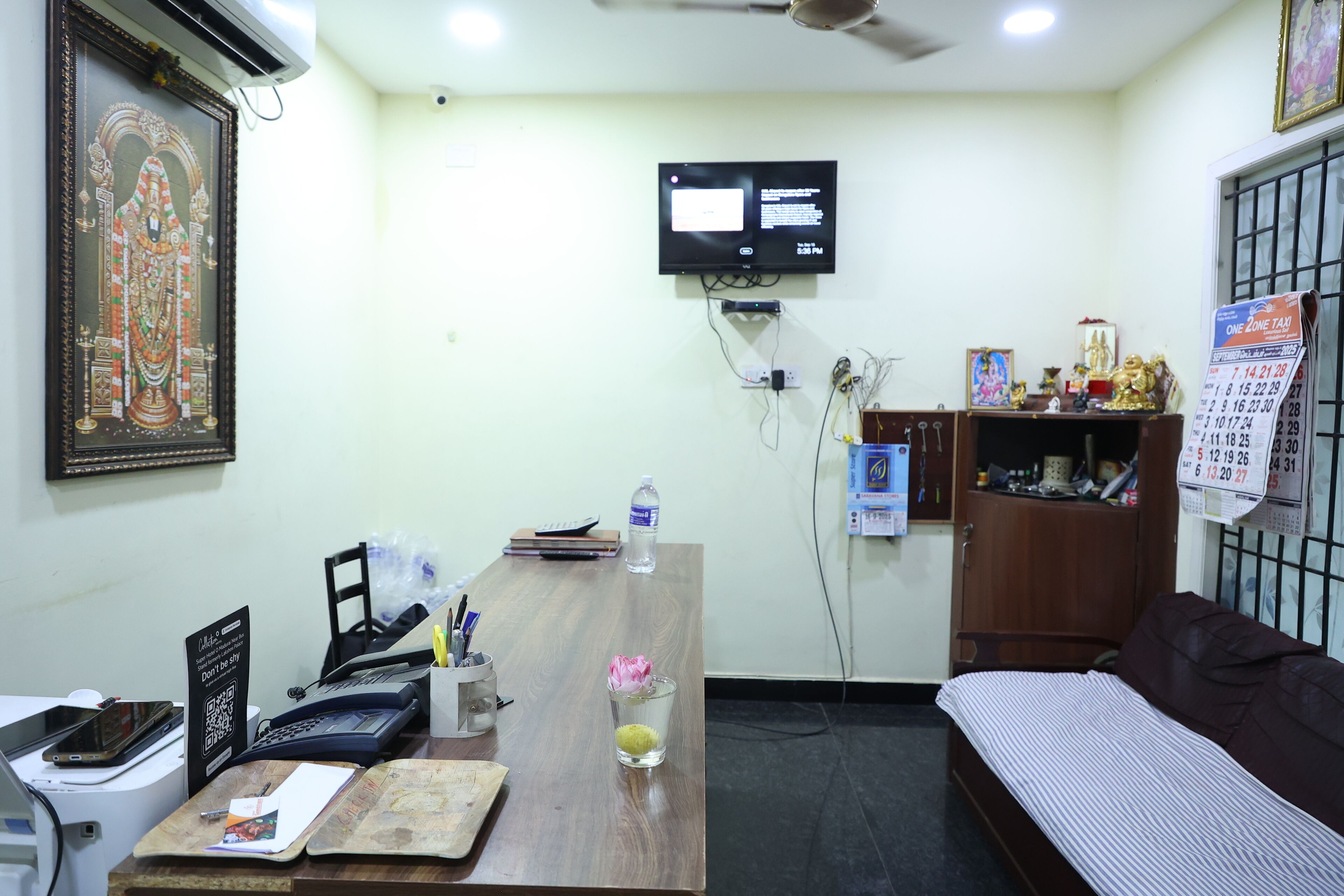 A room with a wooden desk, a bed, a TV mounted on the wall, and a calendar on the wall.