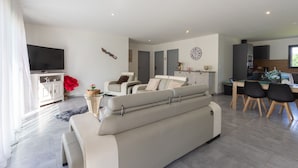 Een moderne woonkamer met een bank, een eethoek met stoelen, een keuken met kastjes, en een aan de muur bevestigde televisie.