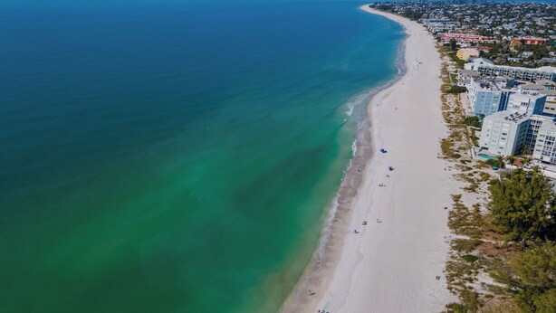 An aerial view of a beach with a long stretch of sand, a clear blue ocean, and a row of buildings along the shore. - Close to beach w/ direct beach access, private fenced yard, Games and beach gear (Holmes Beach)