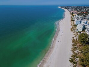An aerial view of a beach with a long stretch of sand, a clear blue ocean, and a row of buildings along the shore. - Close to beach w/ direct beach access, private fenced yard, Games and beach gear (Holmes Beach)