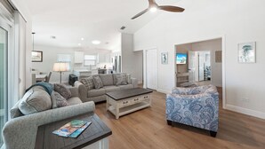 A spacious living room with a sofa, coffee table, and patterned armchair. A ceiling fan is visible, and the room has hardwood flooring. - Close to beach w/ direct beach access, private fenced yard, Games and beach gear (Holmes Beach)